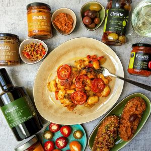 Griechische dicke Bohnen in Tomate-Thymian Sauce auf einem Teller, umgeben von Mezedes wie Oliven, gefüllten Peperoni, Oliven Bruschetta und Produkten von Athanasio Gourmet und Elearte by Athanasio.