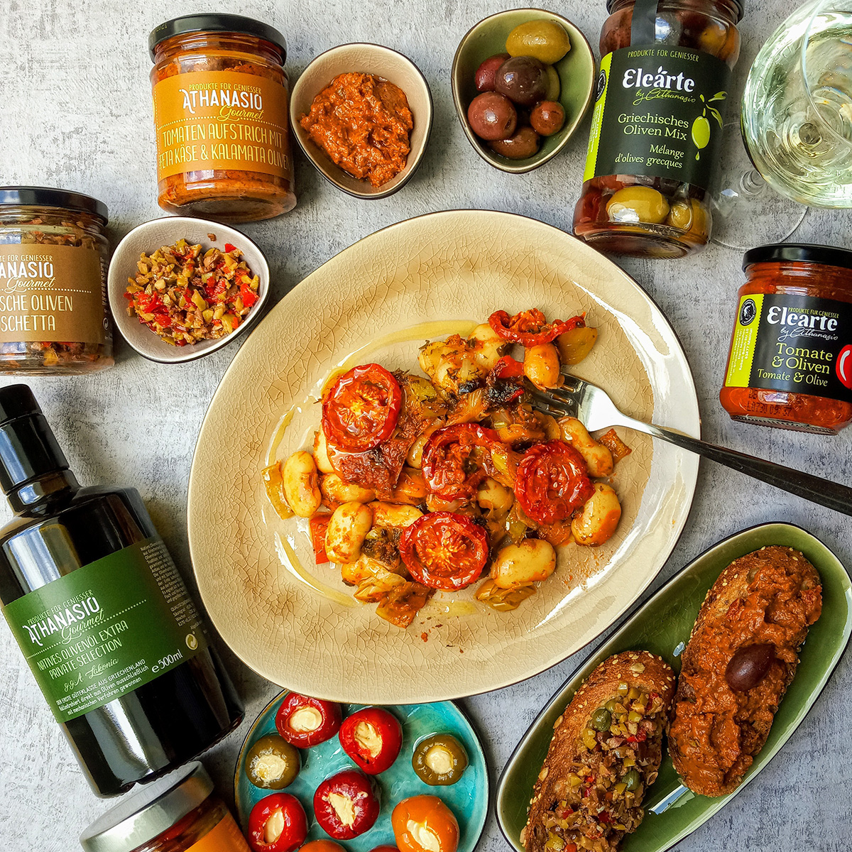 Griechische dicke Bohnen in Tomate-Thymian Sauce auf einem Teller, umgeben von Mezedes wie Oliven, gefüllten Peperoni, Oliven Bruschetta und Produkten von Athanasio Gourmet und Elearte by Athanasio.