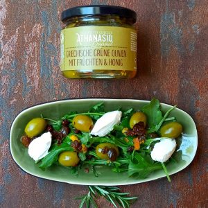 Grüner Salat mit Rucola, Ziegenkäse und griechischen grünen Oliven mit Früchten und Honig von ATHANASIO Gourmet, angerichtet auf Teller neben dem Produktglas.